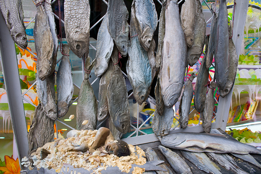 Dead, dried fish with open mouths hang from white iron, attached with red thread. Behind them is a bakery selling neon-frosted cakes and various brightly-colored dry goods. Below the fish, is a blueish, grayish dead man. He looks like he has been mummified. He is lying on a bed of crushed seashells.