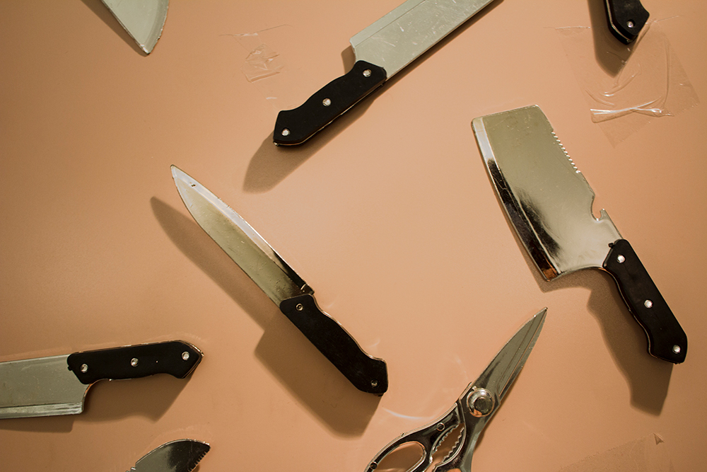 On a flat, peach background, are several different knives. Only some are fully visible: a paring knife, a cleaver, and a pair of scissors. The steel portion of the knives and scissor has been painted silver. There are pieces of transparent tape on parts of the peach surface.