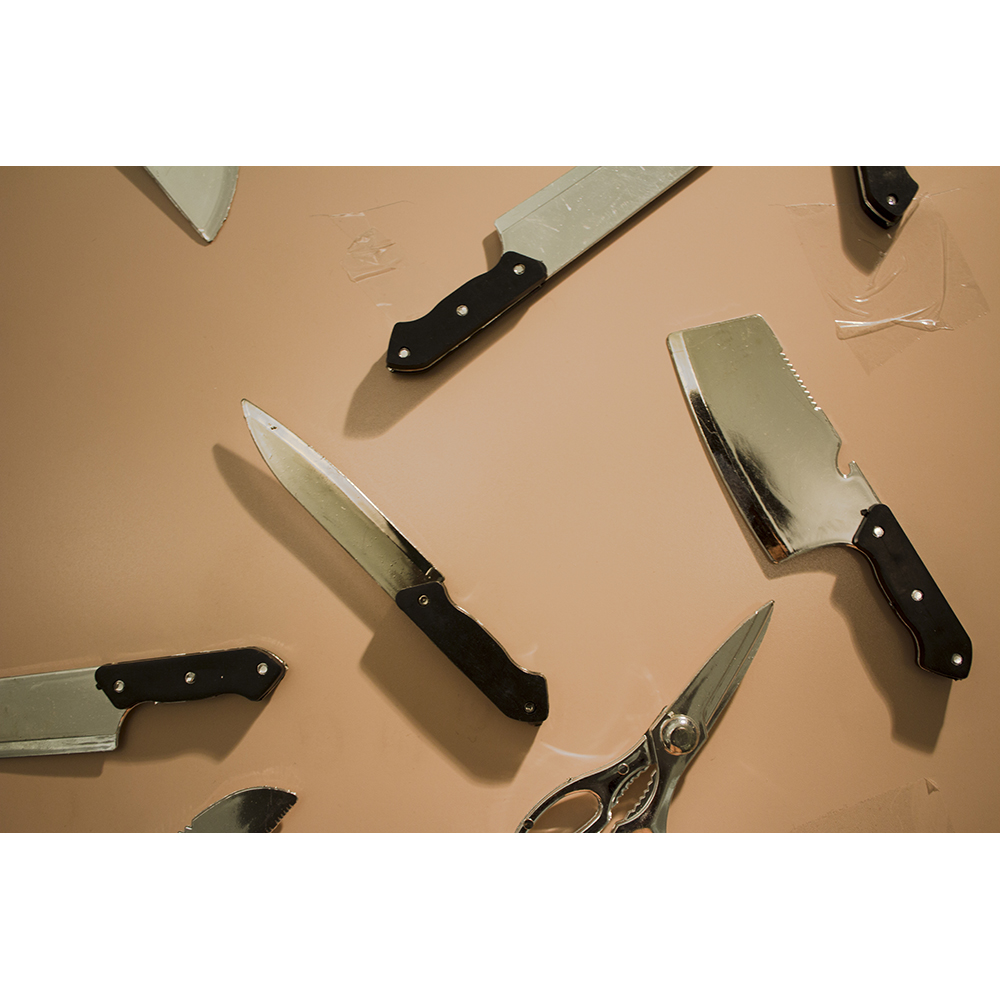 Against a peach-colored surface, several sharp objects are arranged in such a way that they are evenly spaced out from one another. To the left are kitchen knives. At the bottom is a pair of silver scissors. There is also a cleaver. Some of these sharp objects’ handles are taped to the surface with clear packing tape.