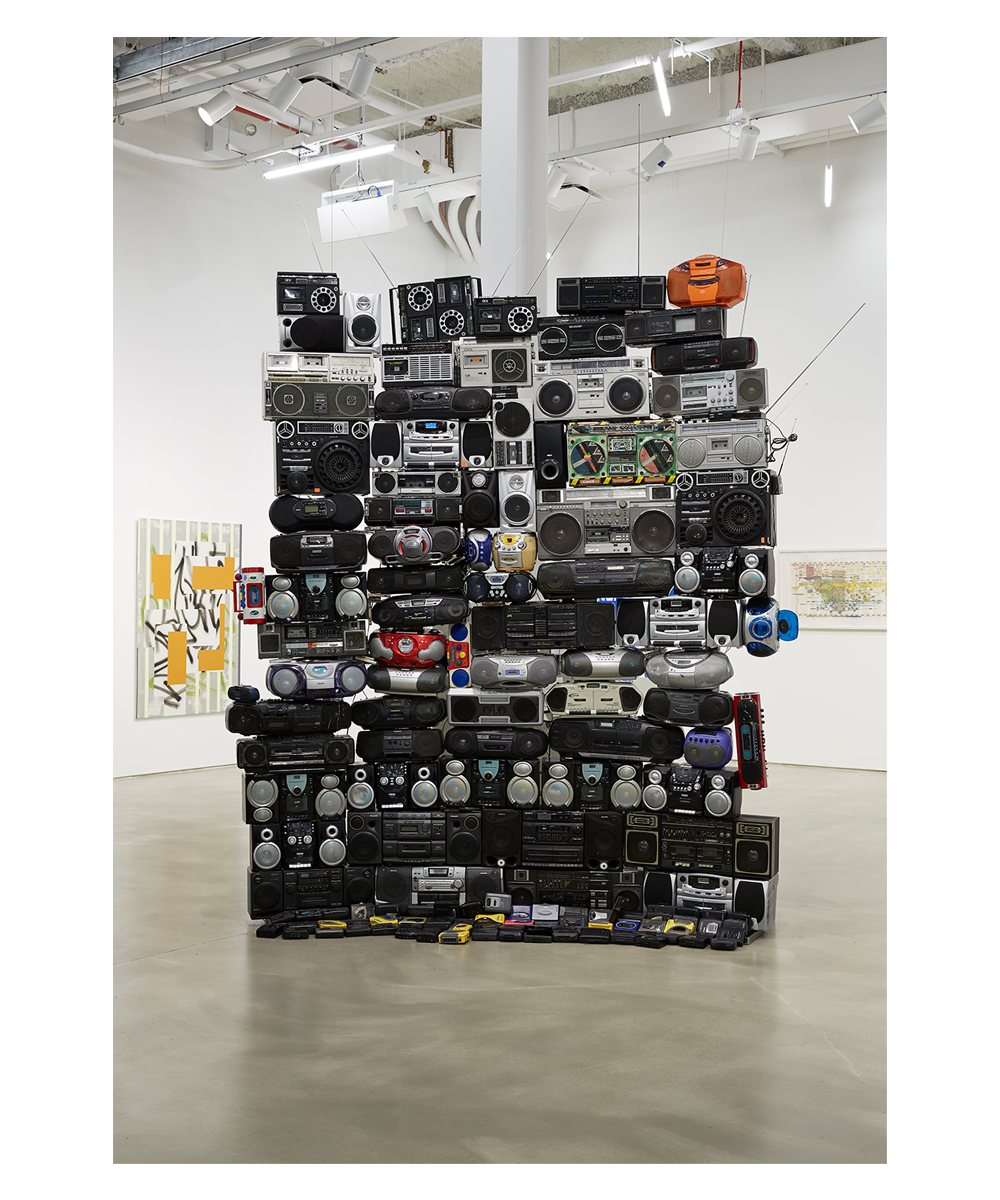 A stack of boomboxes in an art gallery in a neat tight shape—five boomboxes wide, and fourteen boomboxes tall. Most of these boomboxes are black, but there are more colorful ones there as well and a wide range of eras—ranging from radios to tape players to CD players. At the base of the sculpture is a long row of cassette walkmans. 
