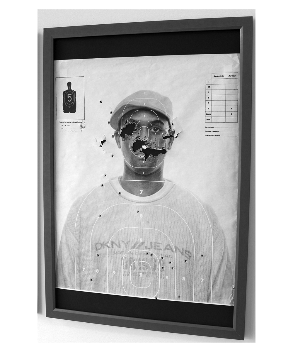 A similar Black-and-white photo of a twenty-something Black man, wearing a DKNY hoodie and Kangol hat, looking directly at the camera, target practice symbols superimposed over his face and chest. He is looking directly at the camera with a neutral expression on his face. Unlike in the first picture,  there are multiple bullet holes in the paper around his head and chest; his left eye, nose, and half of his mouth are almost completely shot off. A thin frame holds the bullet-torn piece of paper.