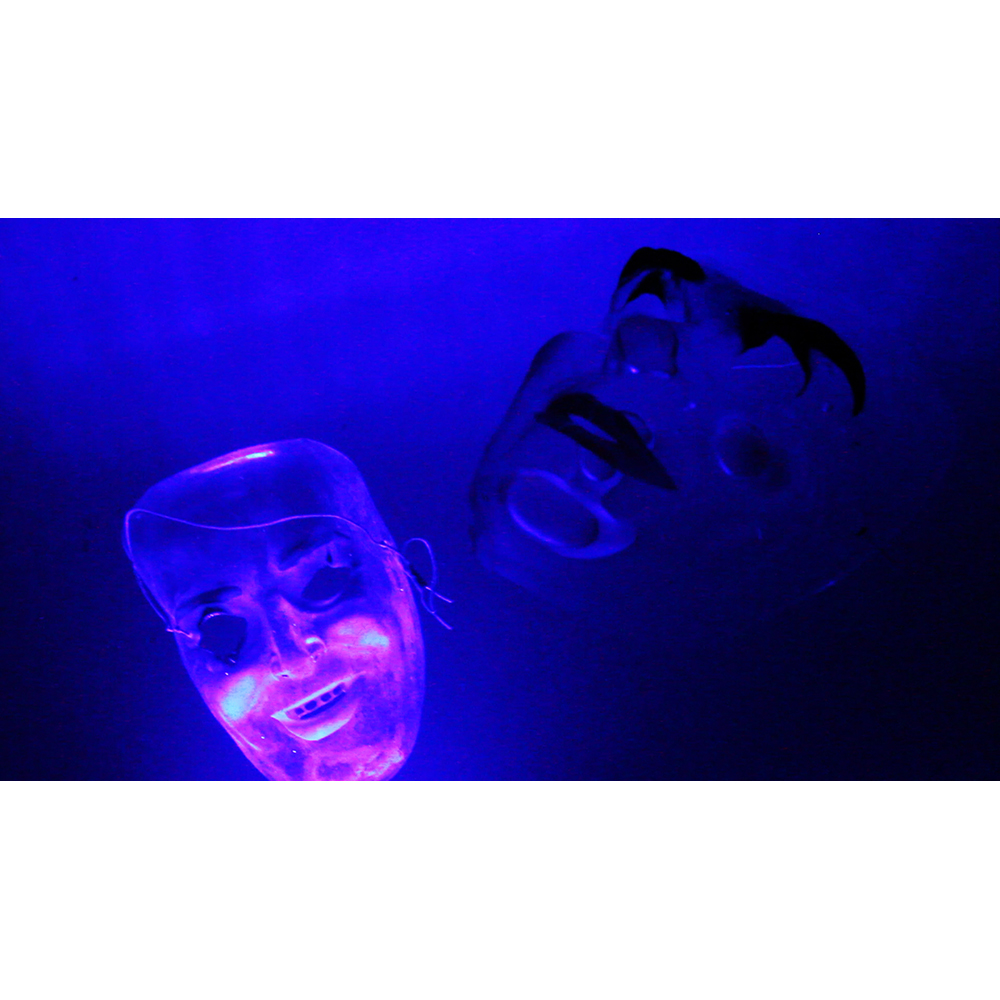 Against a deep blue background, we see two transparent plastic masks, both with smiles plastered across their faces. The one on the left, smaller and further away, is illuminated by bright purple LED’s in its cheeks, coloring the bottom half of the face. The one on the right, much bigger but also more in shadow, has heavy eyebrows and lips painting on, looking almost black in the dim lighting. 