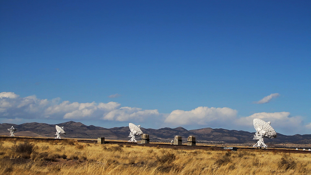 In a big yellow field with dark brown mountains in the backdrop and a bright blue sky, there are four large white satellite dishes in a neat row, each pointed in different directions.