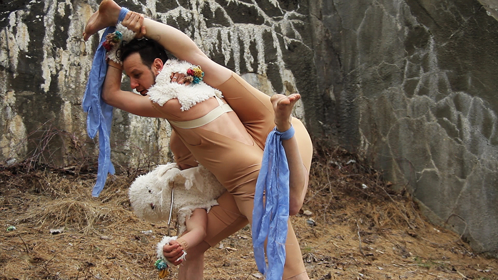 In an area surrounded by gray stone walls are two dancers locked in a contorted pose. One dancer in a beige leotard and white wool collar and cuffs holds another dancer by one leg as they dangle upside down, using the other’s leg to prop themself up at an angle. This dancer wears a white wool mask over their head, concealing their face. Blue scarves are tied to this dangling dancer’s ankles as they are suspended in this pose.