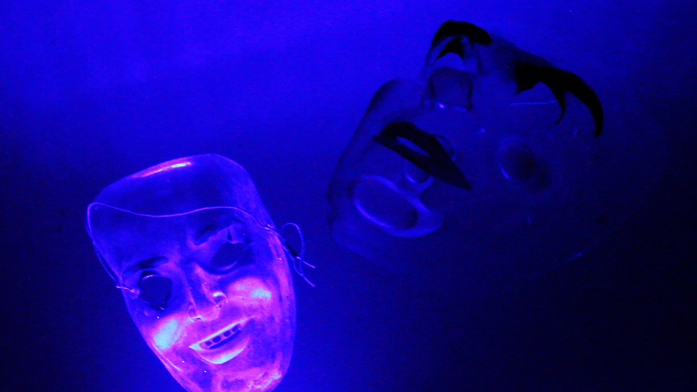 Against an electric blue background are two face masks eerily cast in pink and dark blue light. One mask has the semblance of a human face with holes for eyes and teeth. Its wire hangs over its surface. The other mask is an overlay of two faces, one of which has black eyes and lips peering up, and the other, a stonecut face that is expressionless.