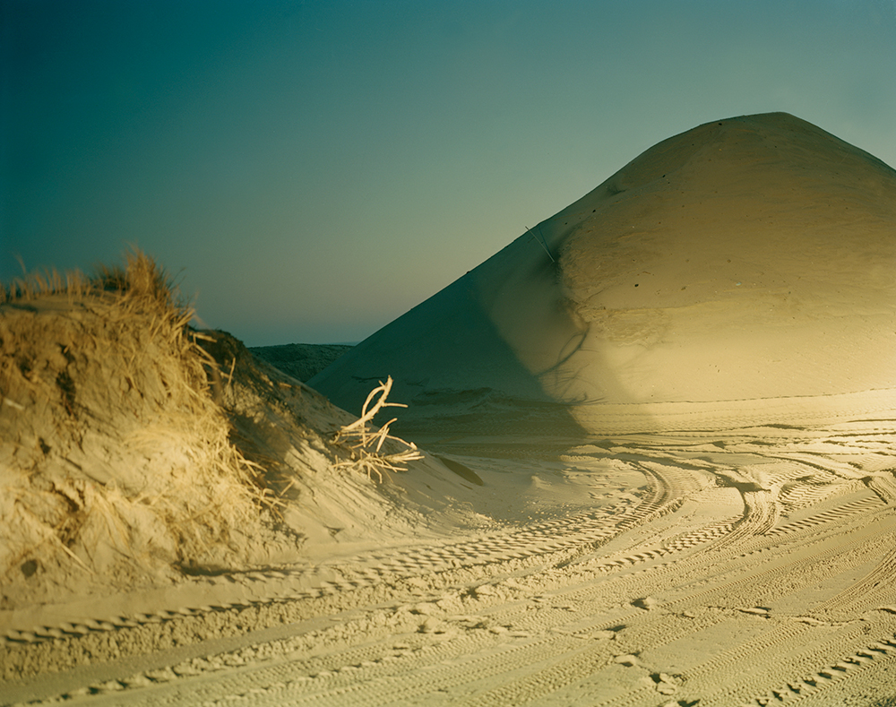 The arrival of dusk in a desert where a dune is prominent. Circling the dune are multiple tire tracks.