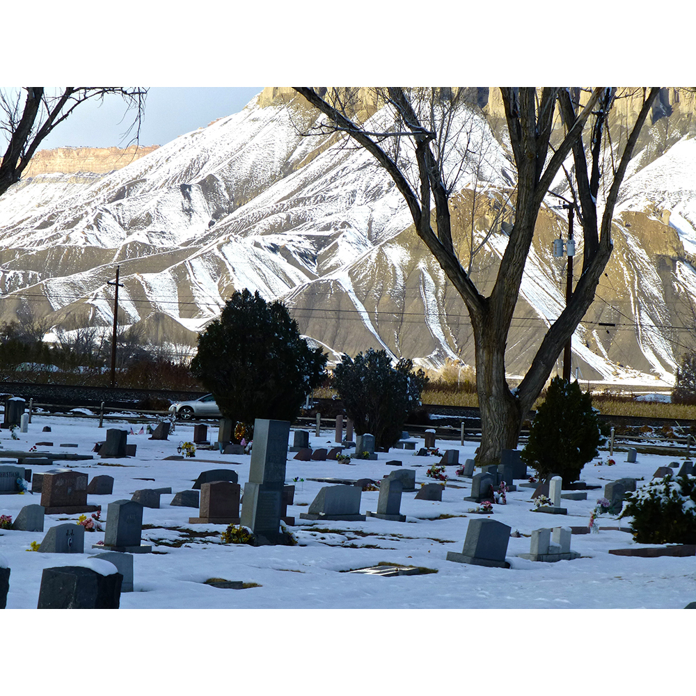 A cemetery slightly covered in snow. In the background, a snow-covered mountain and telephone poles and their wires. 