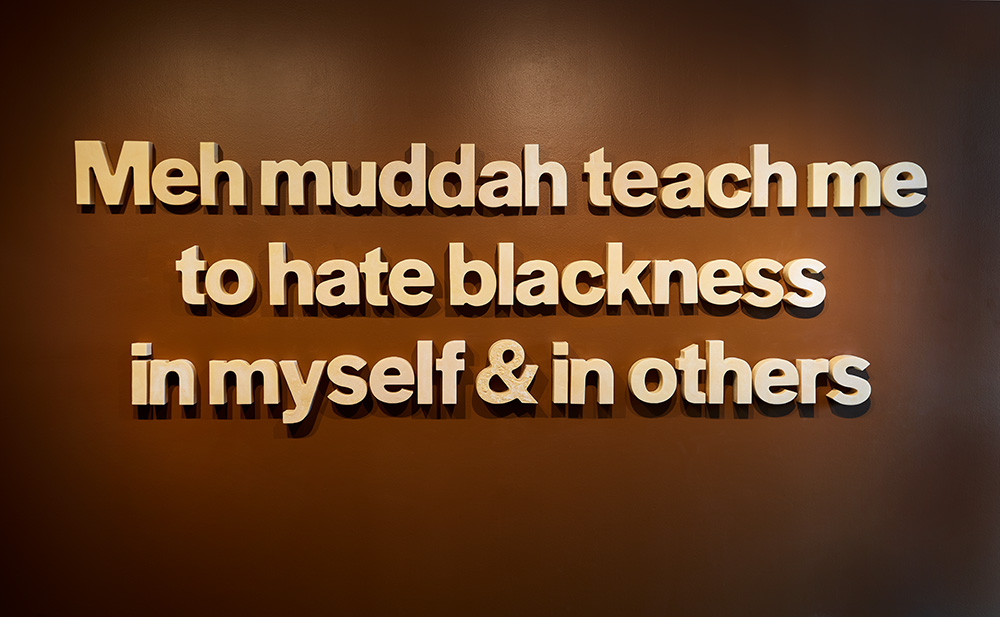 A photograph of an art installation depicting a text-based sculpture made with unrefined cocoa butter and beeswax. The wall is painted a rich, dark brown, and the text, in beige, states “Meh muddah teach me to hate blackness in myself & in others.” 