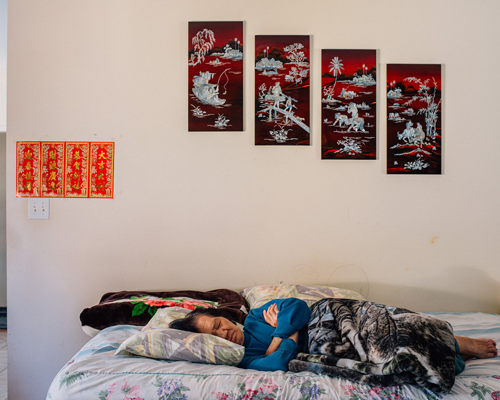 A photograph depicting a room with a bed, folded blankets, and two different pieces of art on the wall. On the bed, an older woman is sleeping. Her face and body face the camera, and her arms are crossed. She’s wearing a blue sweater and a blanket covers her legs, her feet poking out. 