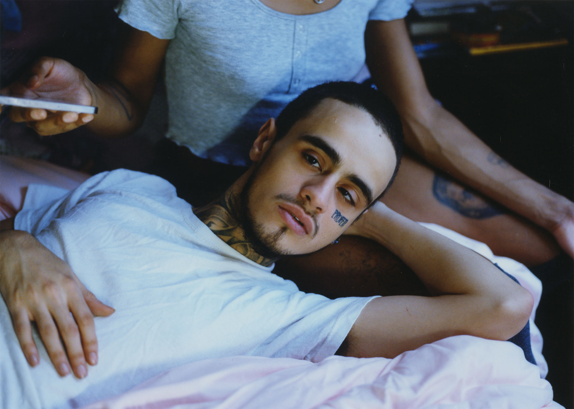 A photograph of a light brown skinned man lying down on a bed. He has one arm draped over his chest, the other arm behind his head. He has a tattoo on his cheek that reads, Prophet. His head is resting against a person's leg.