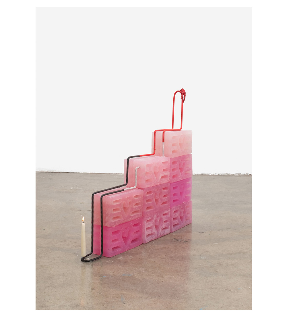 A photograph of a sculpture which is composed of nine bricks painted different shades of pink. The bricks are stacked to resemble stairs. Along the rising brick stairs, there is a thick rope which is black and red, and at the base of the stairs the rope is a candle, which is lit.