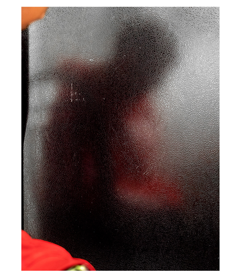 A photograph of frosted glass, behind which there is a blurry figure of a person's body leaning over. There's red fabric in the left-hand side of the image, in front of the glass, and behind the glass the person seems to be wearing red, too.