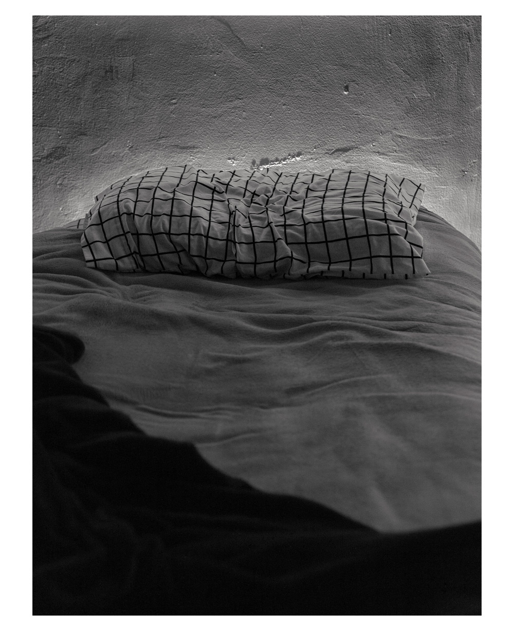 A photograph of an unmade single bed. The photograph is in black and white, and the bedsheets are gray. The pillow in the center of the photograph is white with black checkers. The bed is against a white/gray wall.