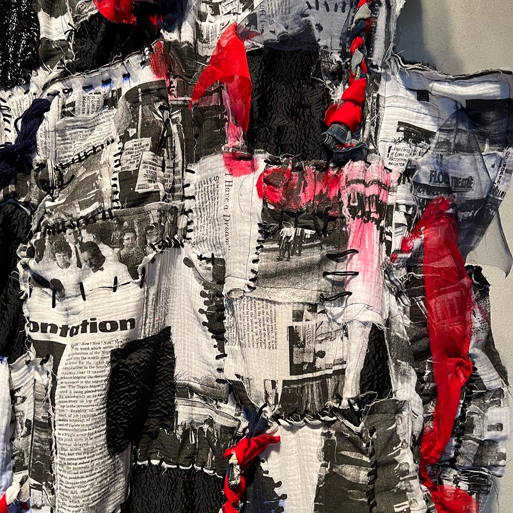 Close up of long black white and red tapestry, shows news articles printed on fabric stitched together.