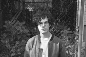 A black and white photo of the poet with glasses, a white t-shirt, a jacket, and short curly hair, standing in front of a chain link fence and some plants.