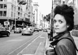 The poet stands on the right-side edge of the black and white photo with an up-do hairstyle wearing a nose-ring, a blazer, and a backpack, looking toward the viewer, in front of a NYC street-scene of buildings, people, and cars.